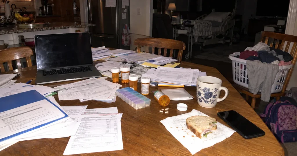 A kitchen table at night covered in bills and medical forms, pill bottles, a pill organizer and a half-eaten sandwich, with a hospital bed in the dim room beyond.