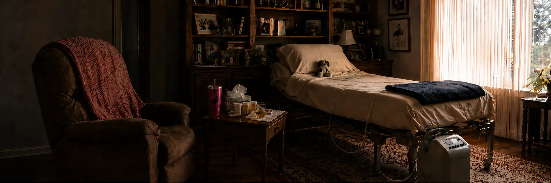 A hospital bed set up in a book-lined living room, with a recliner, throws, medications, and an oxygen concentrator nearby.
